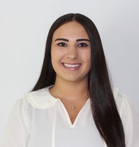 Smiling woman with long dark hair wearing a white blouse, shown from mid-face to chest in a pale background