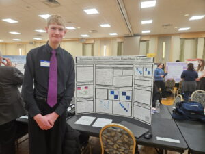 Young man in a black shirt and purple tie stands beside a science-fair poster about algorithmic classification of classical piano music.