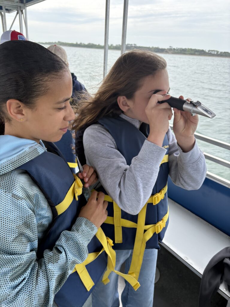 Two people in life jackets on a boat; one girl looks through binoculars toward the water while the other watches nearby.