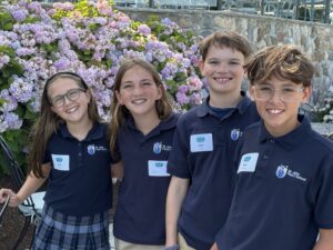Four smiling students in navy polo shirts pose in front of a stone wall and pink/purple hydrangea bushes.