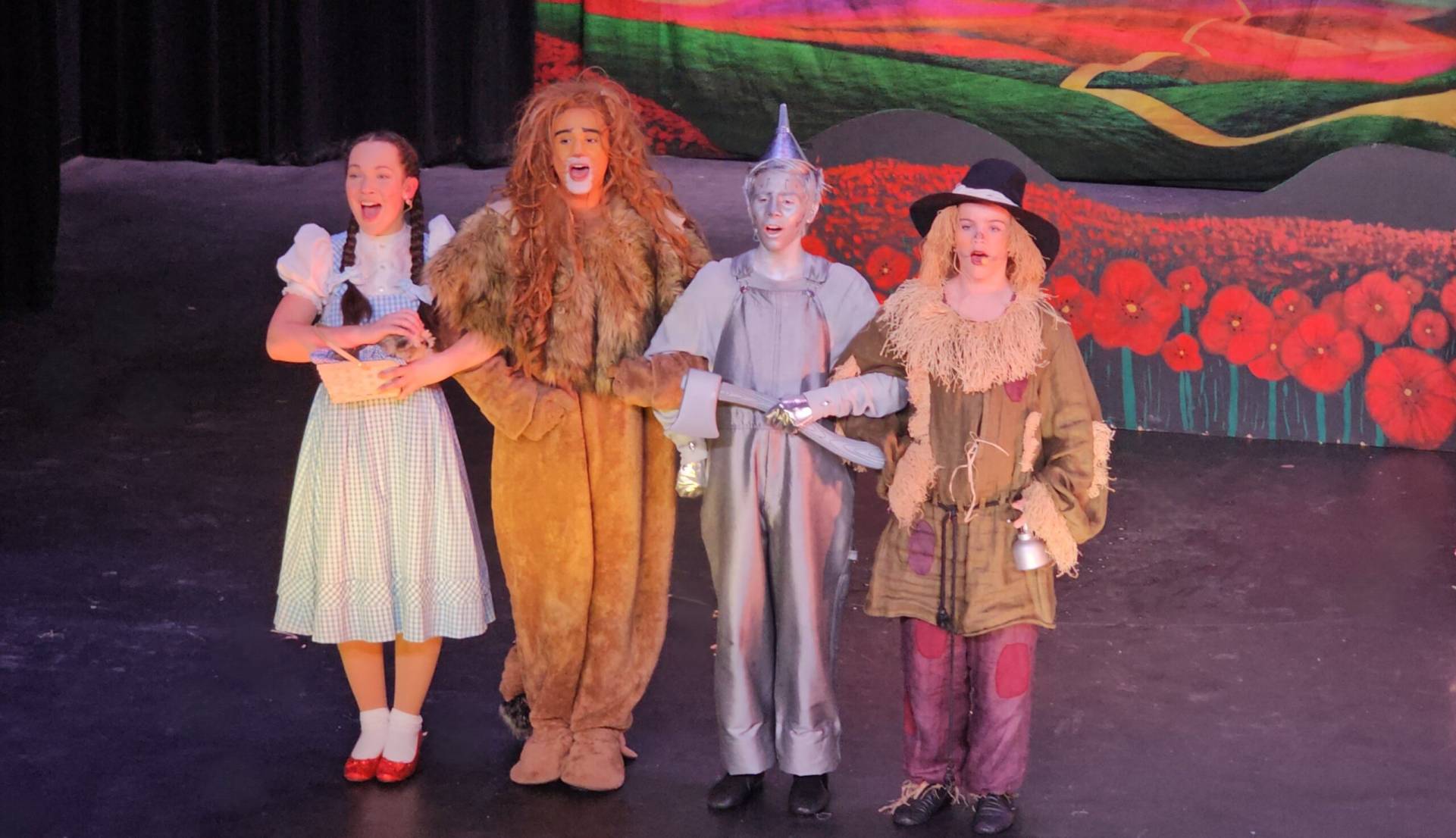 Four children in Oz-inspired costumes stand on a stage: Dorothy with a basket, the Cowardly Lion, Tin Man, and Scarecrow before a bright backdrop of red flowers.